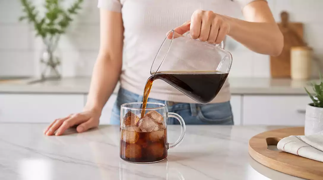 Person pouring cold brew concentrate from a glass pitcher into a mug with ice, natural light, kitchen background